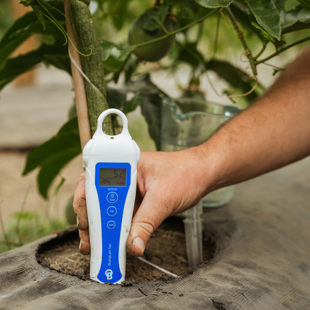 A hand holding a white and blue digital soil moisture meter next to a plant in a garden bed, with soil, green leaves, and a watering can visible in the background.