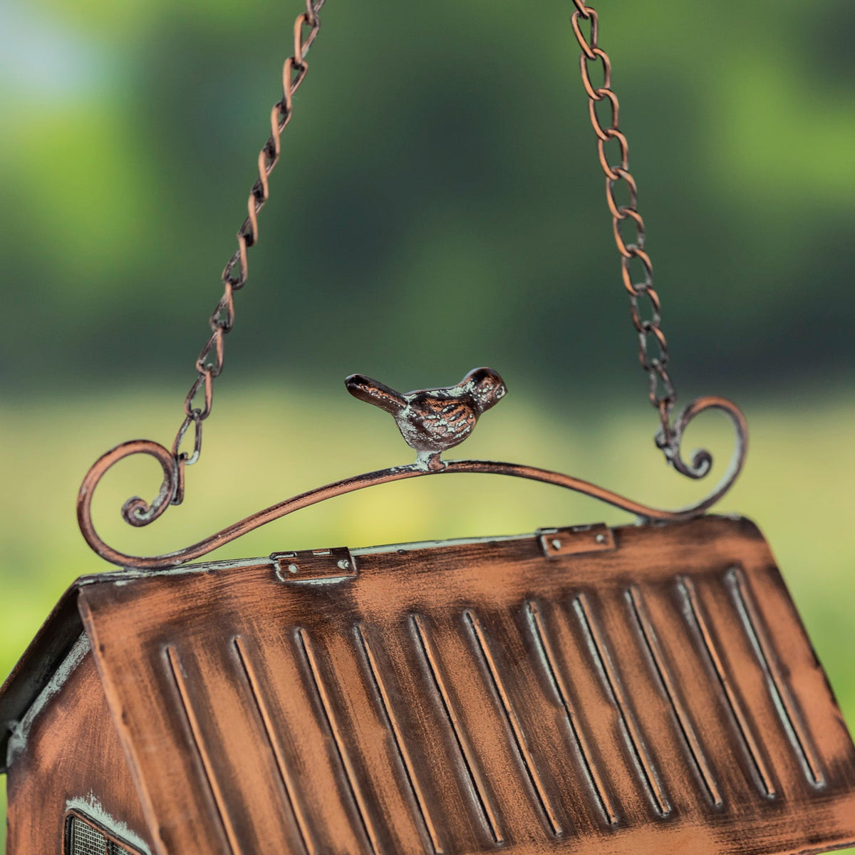 A close-up of a hanging birdhouse ornament with a small bird figure on the top bar, suspended by chains against a blurred green outdoor background.