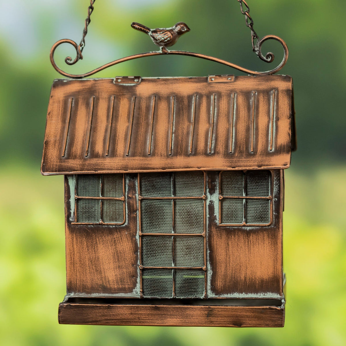 A rustic metal bird feeder shaped like a small house with a slatted roof, mesh windows, and a hanging chain, shown against a blurred green outdoor background.