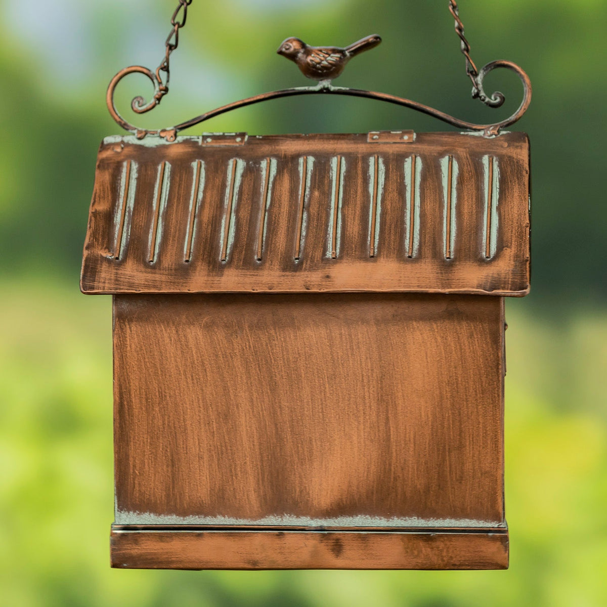 A rustic copper bird feeder with a textured, weathered finish and a small bird figurine on the top bar, shown hanging against a soft green bokeh background.