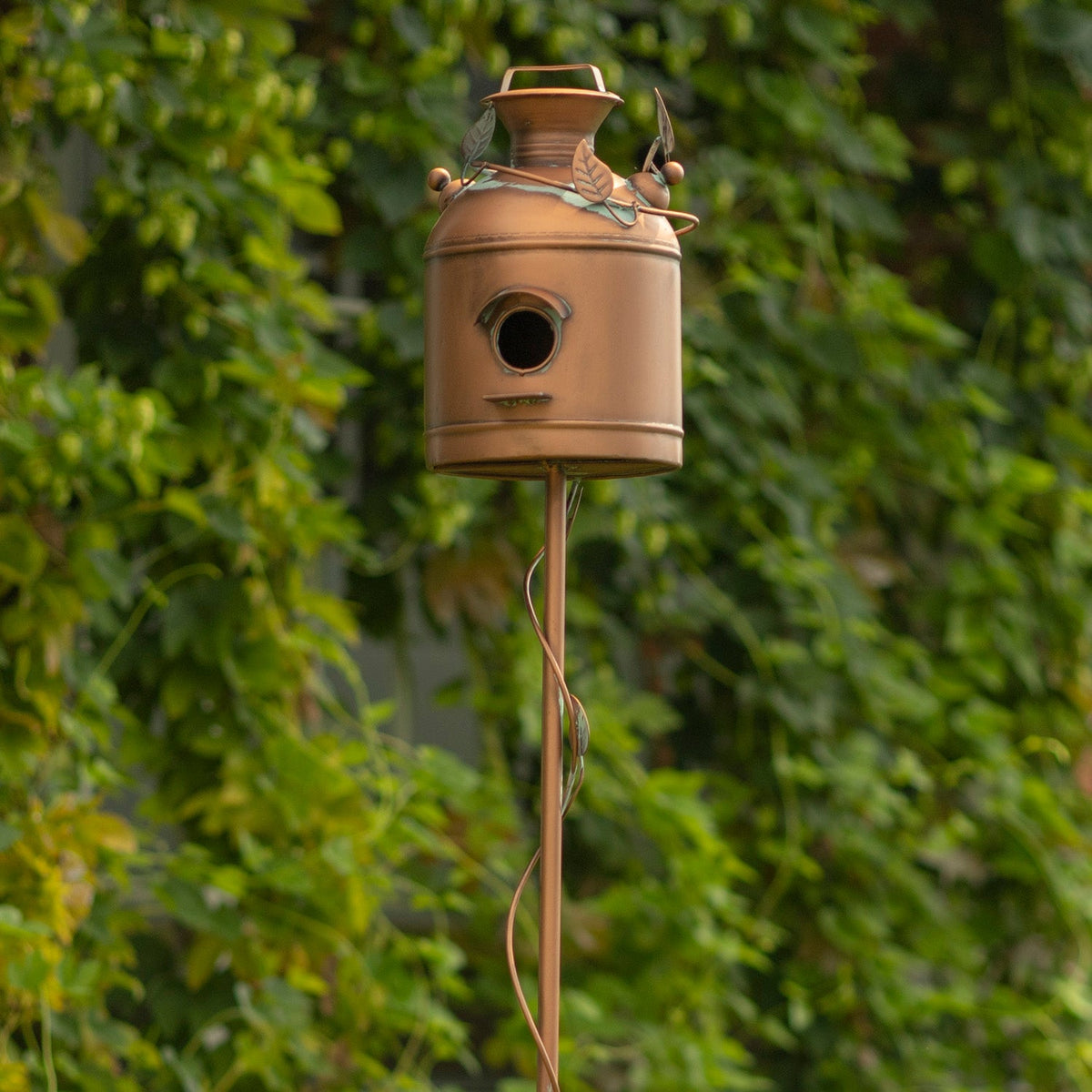 A rustic metal birdhouse shaped like a vintage milk canister, mounted on a tall matching pole, set against a background of green foliage.