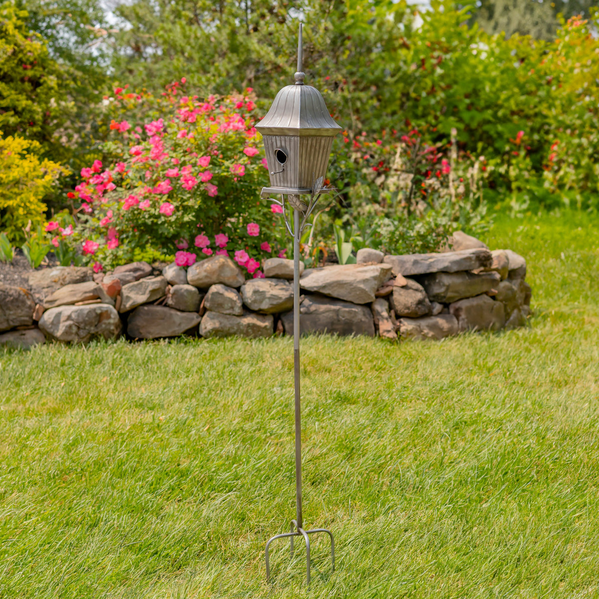 A metal garden birdhouse on a tall stake with a three-pronged base, set on a green lawn in front of a stone wall and flowering bushes.