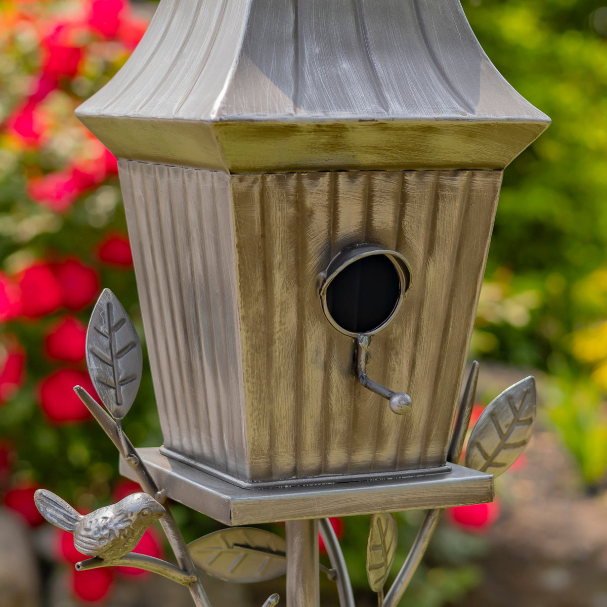 A close-up of a metal birdhouse with a weathered silver and gold finish, mounted on a decorative branch-and-bird stand in a garden setting with blurred red flowers and green foliage.