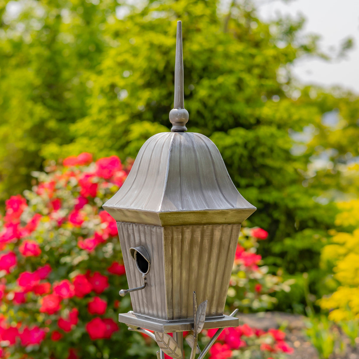 A metal birdhouse with a weathered silver-gray finish and a pointed finial, set against a blurred garden background of green foliage and pink flowers.