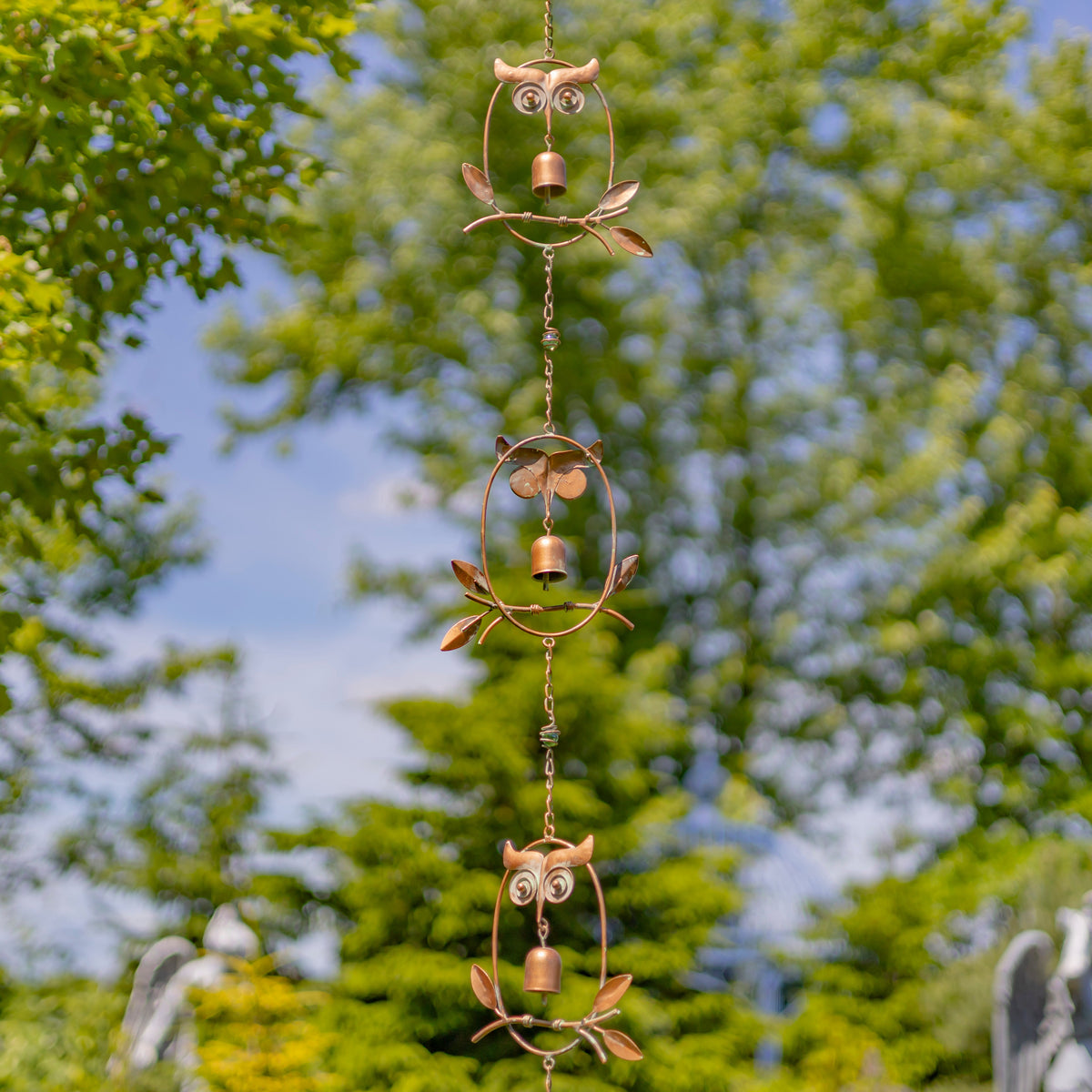 A hanging metal owl wind spinner with three stacked owl figures and small bells, photographed outdoors against a blurred background of green trees and blue sky.