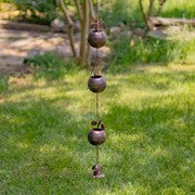 A hanging wind chime with three dark metal bells and a small top striker, photographed outdoors on a grassy lawn with trees in the background.