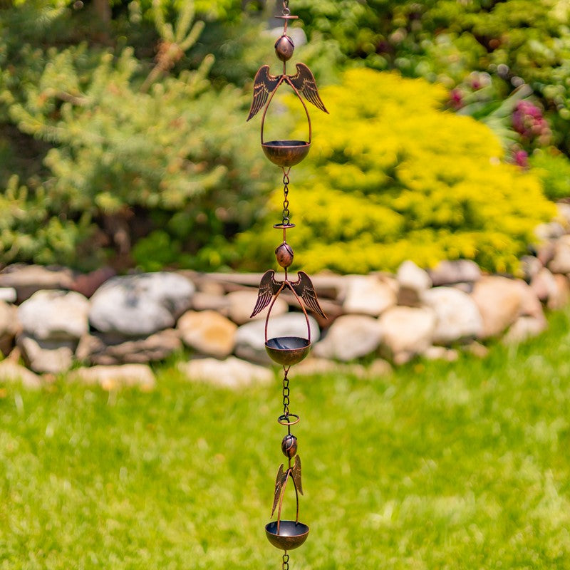 A hanging metal garden wind spinner with angel and bird motifs, photographed outdoors against a blurred green lawn and shrubs.