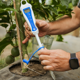 A person in a greenhouse uses a white and blue digital soil moisture meter attached to a clear plastic tube, measuring moisture around a young plant.