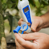 A close-up of a person's hands holding a white and blue digital pH meter and a clear plastic bottle with a blue cap, likely in a garden or greenhouse setting.