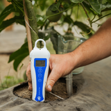 A hand holding a white and blue digital soil moisture meter next to a plant in a garden bed, with soil, green leaves, and a watering can visible in the background.