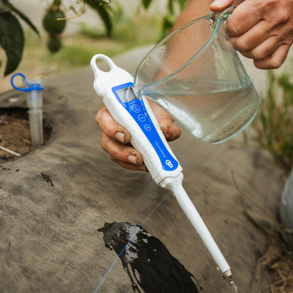 A close-up of a person watering a garden bed using a white and blue electronic drip irrigation controller, pouring water from a clear glass measuring cup.