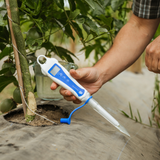 A person's hand holds a white and blue digital soil moisture meter with a probe inserted into the ground next to a potted plant in a garden setting.