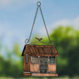 A hanging bird feeder shaped like a small rustic house, with a copper-toned metal body, chain hanger, window details, and a small bird figure on top, set against a blurred outdoor background.