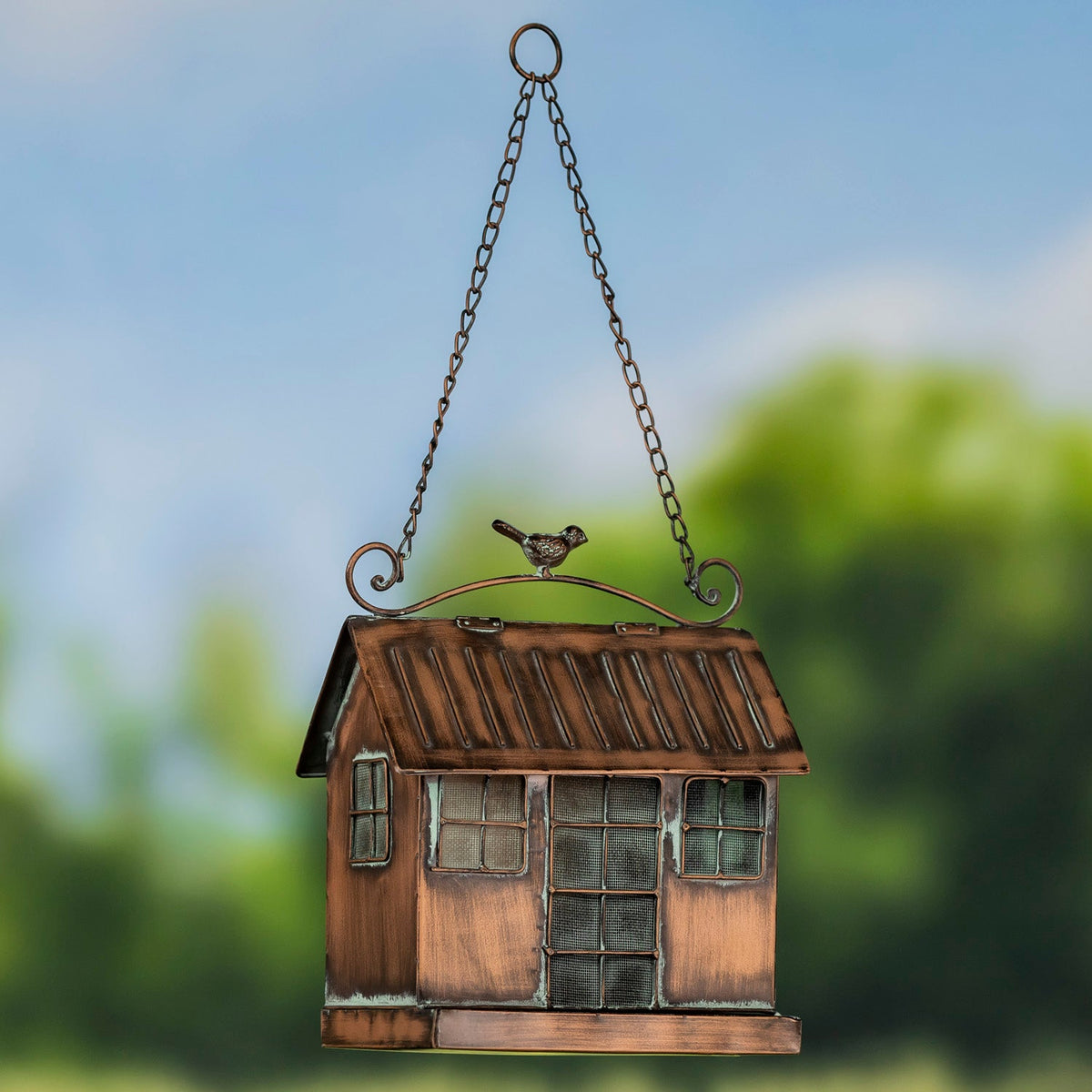A hanging bird feeder shaped like a small rustic house, with a copper-toned metal body, chain hanger, window details, and a small bird figure on top, set against a blurred outdoor background.