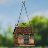 A hanging bird feeder shaped like a small rustic house, with a copper-toned metal body, chain hanger, window details, and a small bird figure on top, set against a blurred outdoor background.