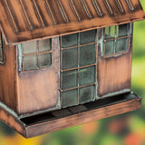A close-up of a small house-shaped bird feeder with a weathered copper-brown finish, mesh screens, and a slatted roof, shown against a blurred outdoor background.