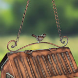 A close-up of a hanging birdhouse ornament with a small bird figure on the top bar, suspended by chains against a blurred green outdoor background.