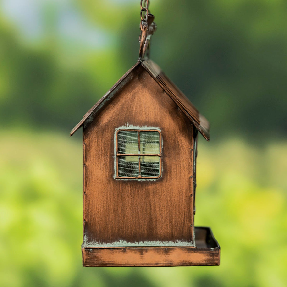 A small rustic wooden birdhouse feeder hanging from a metal chain, with a weathered brown finish and a small window on the front, set against a soft green bokeh background.