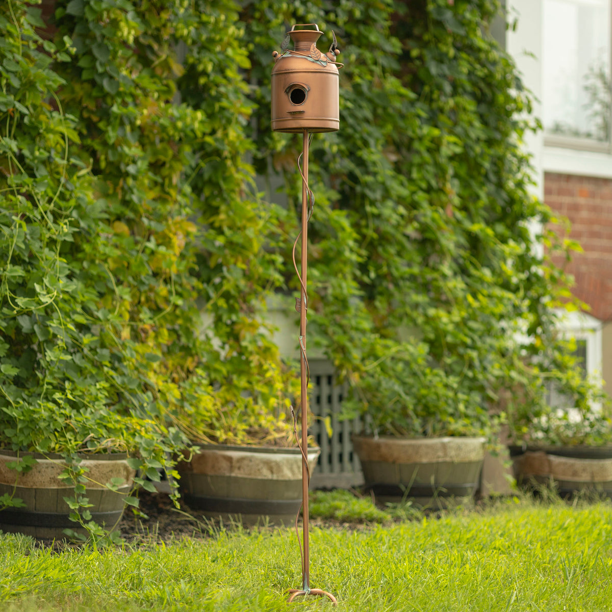 A rustic copper birdhouse on a tall twisted metal stake, set in a grassy garden with a leafy green houseplant background.