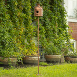 A rustic copper birdhouse on a tall twisted metal stake, set in a grassy garden with a leafy green houseplant background.