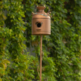 A rustic metal birdhouse shaped like a vintage milk canister, mounted on a tall matching pole, set against a background of green foliage.