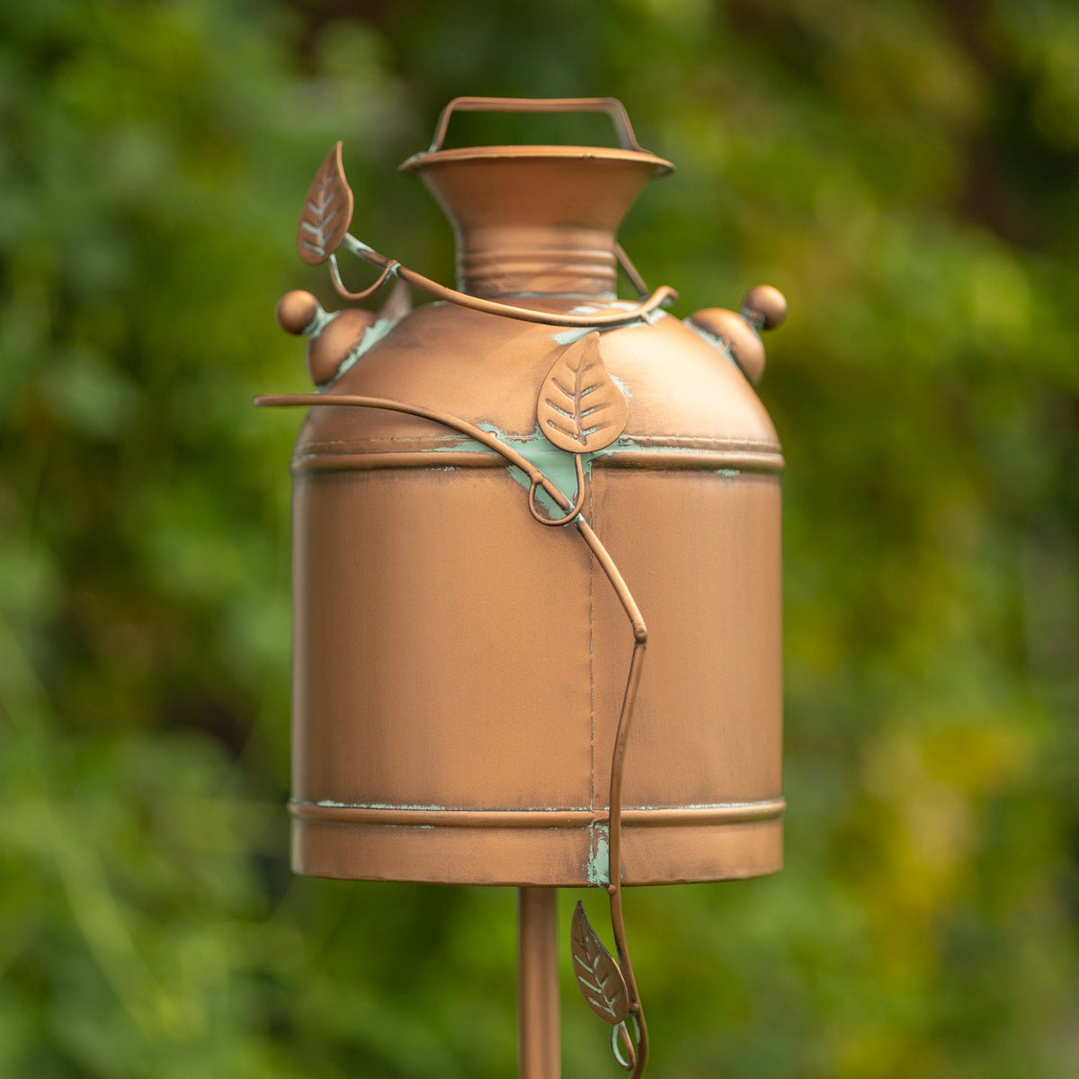 A decorative copper-toned metal garden stake shaped like a vintage milk canister, with leaf-shaped accents and a small top handle, photographed outdoors against a blurred green foliage background.