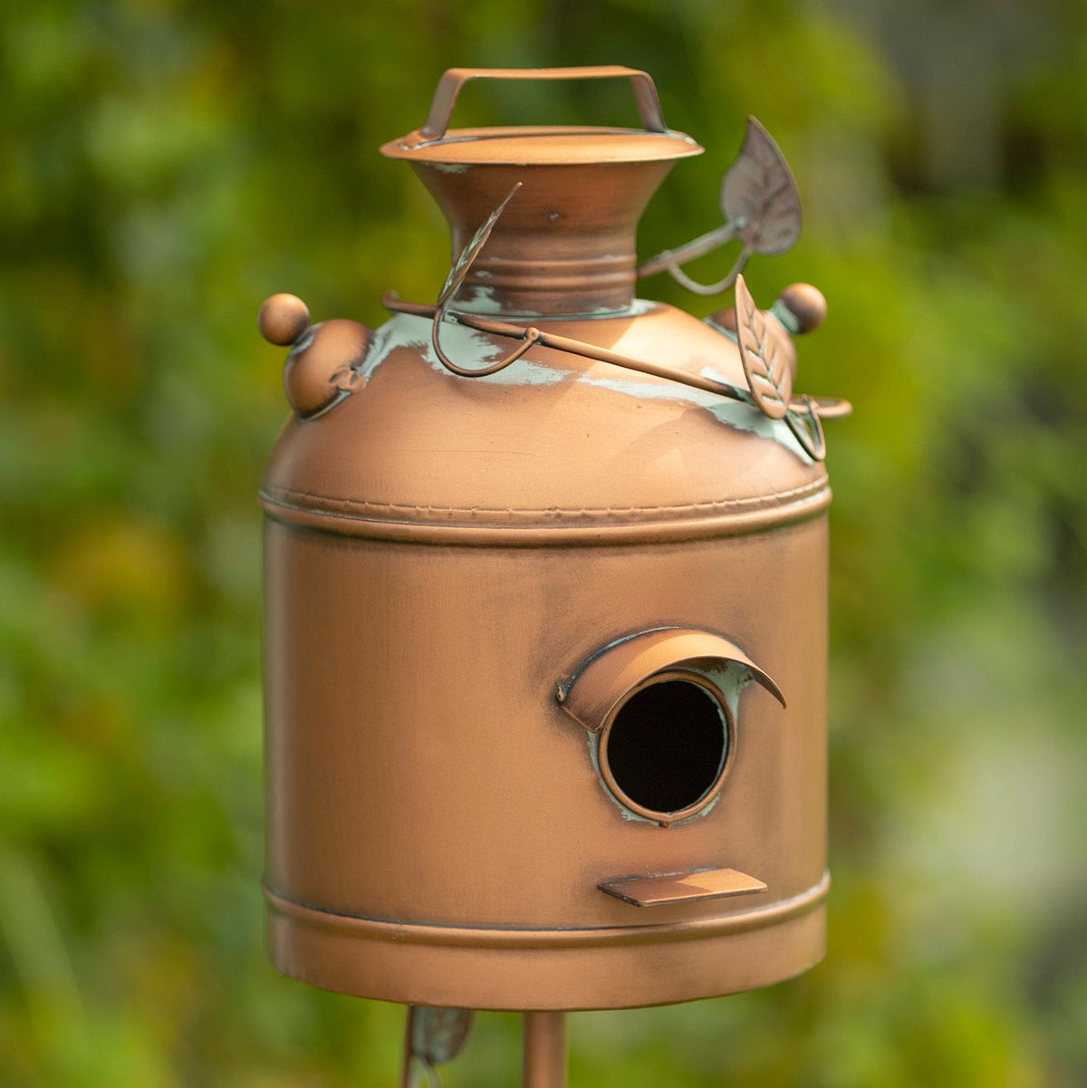 A decorative metal birdhouse shaped like a vintage milk canister, with a copper-toned finish, leaf and vine accents, and a small round entrance hole, photographed outdoors against a blurred green garden background.