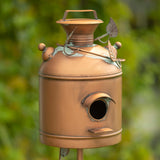 A decorative metal birdhouse shaped like a vintage milk canister, with a copper-toned finish, leaf and vine accents, and a small round entrance hole, photographed outdoors against a blurred green garden background.