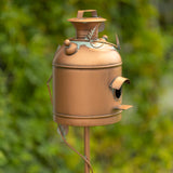 A rustic copper birdhouse shaped like a vintage milk canister, mounted on a metal stake against a blurred green garden background.