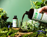Hand pouring a liquid from a bottle labeled 'REV' into a glass with plants and flowers in the background.