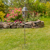 A metal garden birdhouse on a tall stake with a three-pronged base, set on a green lawn in front of a stone wall and flowering bushes.