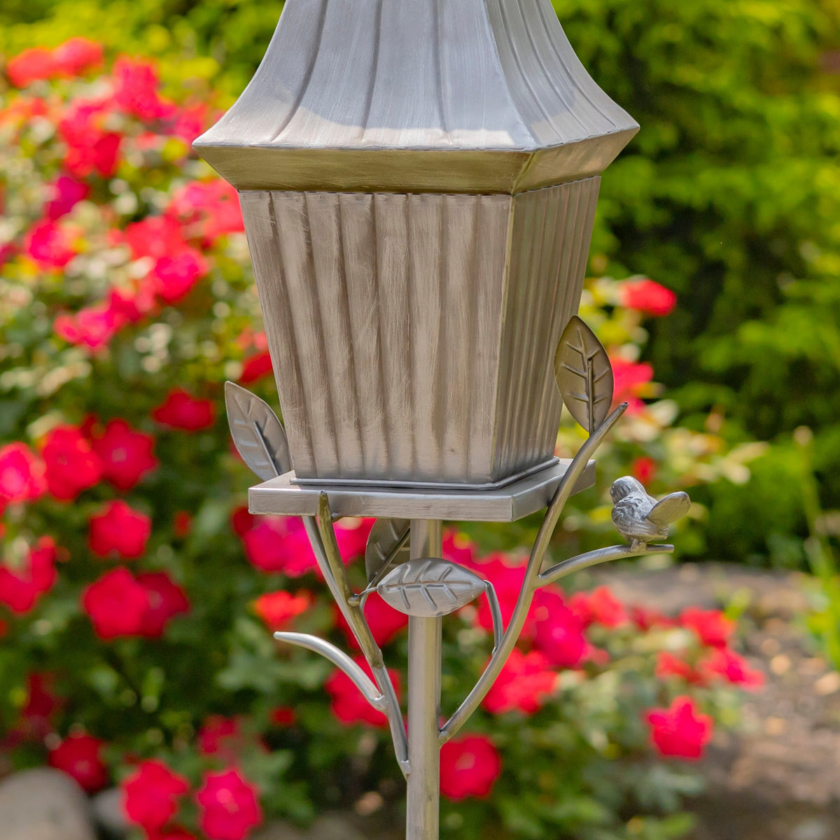 Decorative bird feeder with a bird on a stand in front of pink flowers