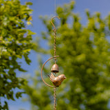 A small copper bird wind chime hanging outdoors against a blurred background of green trees and blue sky.