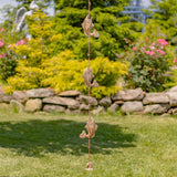A vertical metal wind spinner featuring three dangling fish charms and a small bird at the bottom, displayed outdoors on a green lawn with a stone wall and garden foliage in the background.
