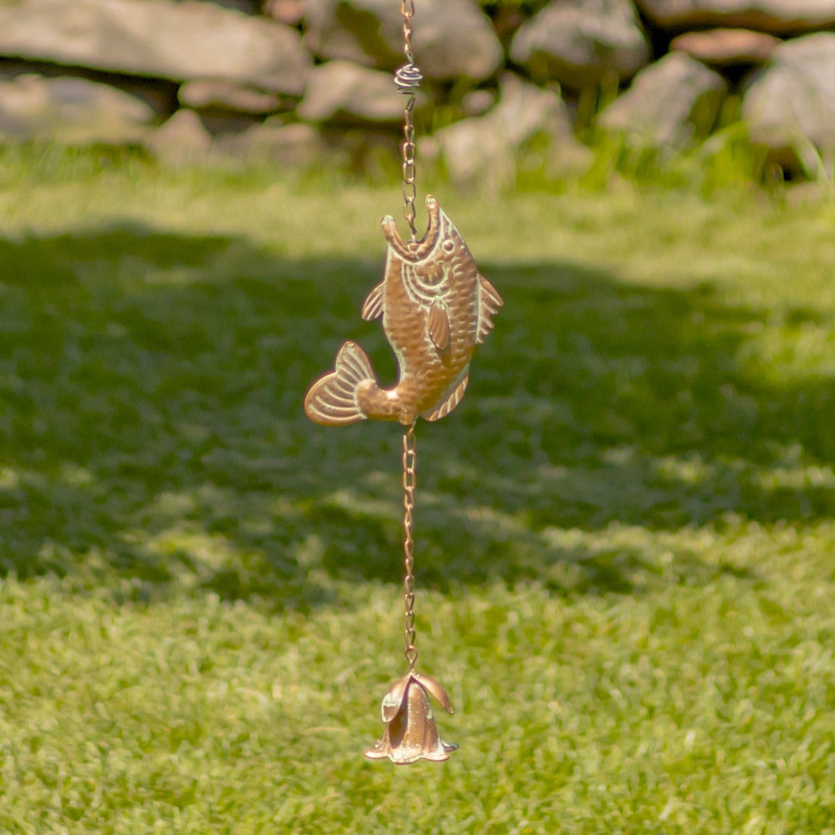 Decorative fish-shaped wind chime hanging on grass with a stone wall in the background
