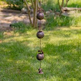 A hanging wind chime with three dark metal bells and a small top striker, photographed outdoors on a grassy lawn with trees in the background.